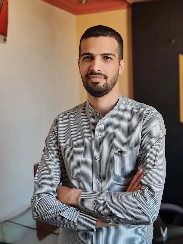 Mohamed Tanash standing indoors with his arms crossed. He has short dark hair, a trimmed beard, and is wearing a light-colored, vertically striped shirt with buttons. The background features a neutral wall and some subtle elements of furniture and decor, such as a chair and a corner with contrasting colors. The lighting is soft and natural.